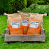 Three bags of Ark brand bird food placed in a wooden crate on a grassy lawn. The bags are labeled as 'Ark Wildlife Premium Wild Bird Food', 'Ark Wildlife Premium Songbird Food', and 'Ark Wildlife Hearty Mix'.