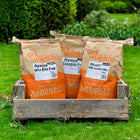 Three bags of Ark brand bird food placed in a wooden crate on a grassy lawn. The bags are labeled as 'Ark Wildlife Premium Wild Bird Food', 'Ark Wildlife Premium Songbird Food', and 'Ark Wildlife Hearty Mix'.