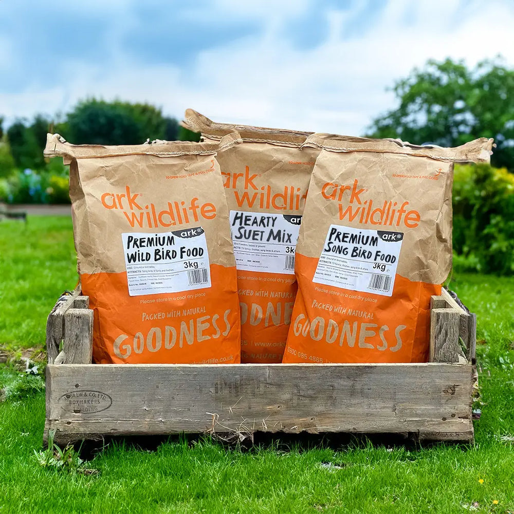 Three bags of Ark Wildlife bird food in a wooden crate on grass with trees in the background