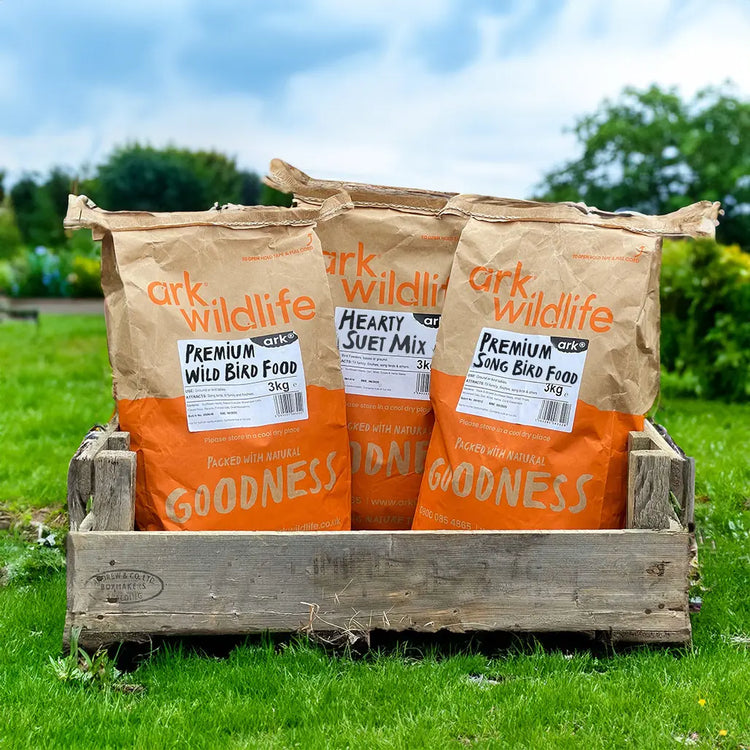 Three bags of Ark Wildlife bird food in a wooden crate on grass with trees in the background