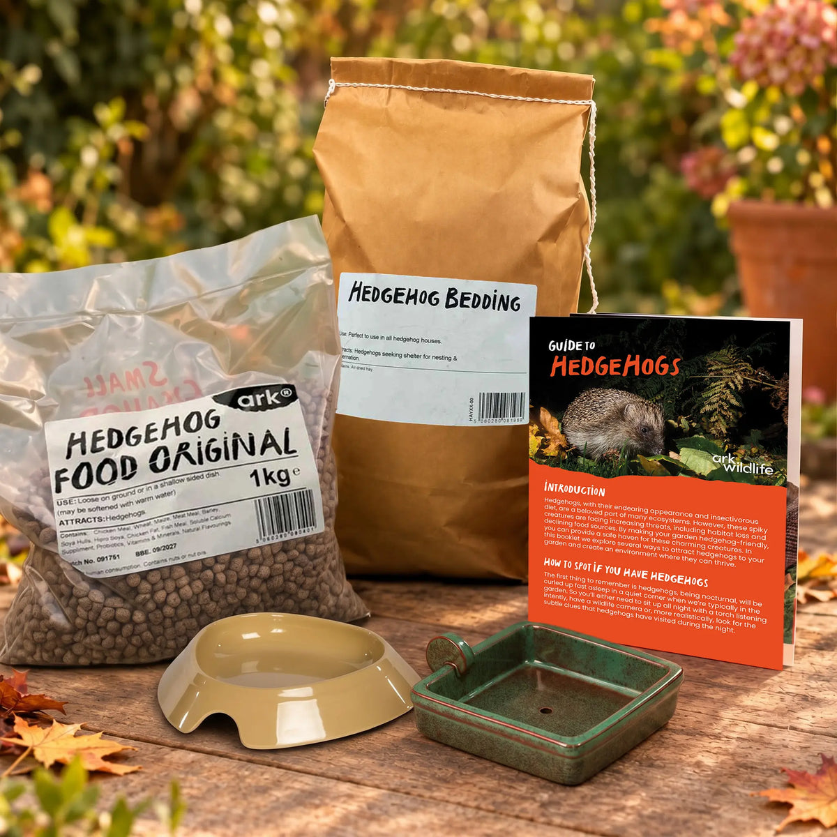 Hedgehog care starter kit displayed on a wooden table outdoors, featuring a bag of Ark Hedgehog Food Original, a paper bag of Hedgehog Bedding, an instruction booklet, a water bowl, and a green food bowl, with a blurred garden background.