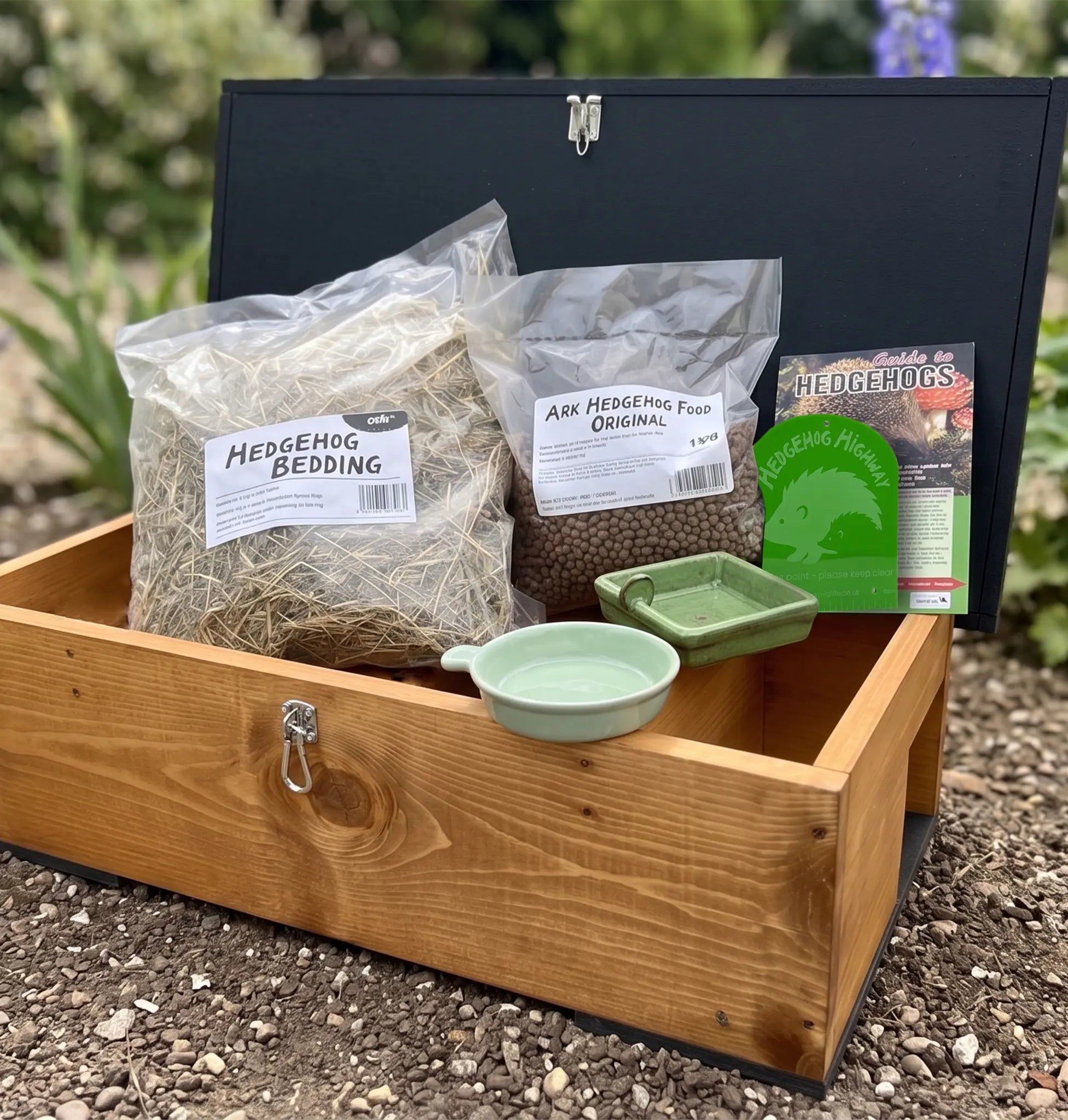 A hedgehog house including bags of hedgehog bedding and food, a green bowl, a small green dish, and a hedgehog bedding card, set outdoors on gravel with blurred greenery in the background.