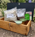 A hedgehog house including bags of hedgehog bedding and food, a green bowl, a small green dish, and a hedgehog bedding card, set outdoors on gravel with blurred greenery in the background.