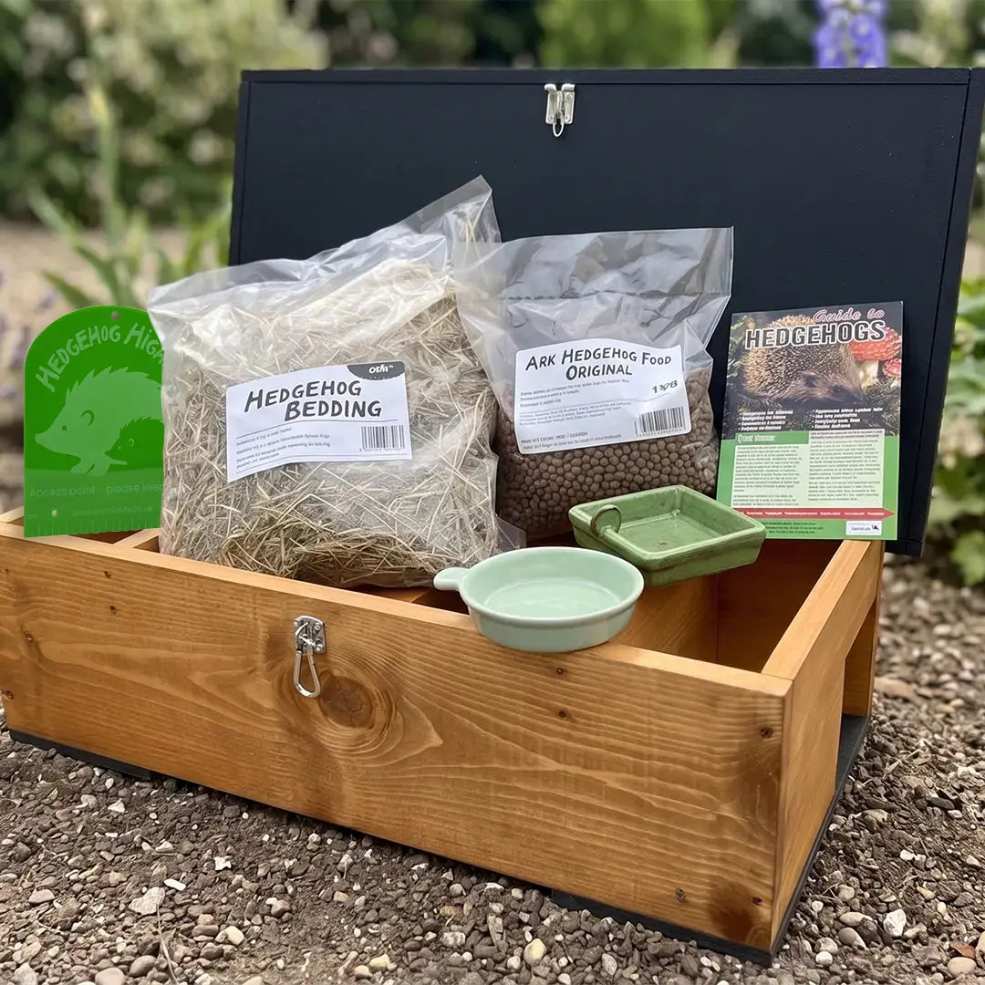 A wooden hedgehog house shown outdoors on gravel with hedgehog bedding, food, bowls, and a hedgehog highway sign included in the set.