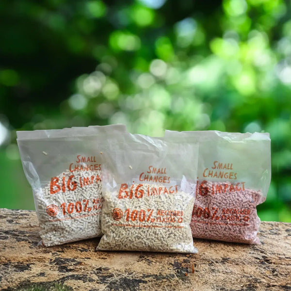 Three clear recyclable plastic bags filled with suet pellets printed with 'Small Changes Big Impact' and '100% Natural', arranged on a stone surface with a blurred green outdoor background.