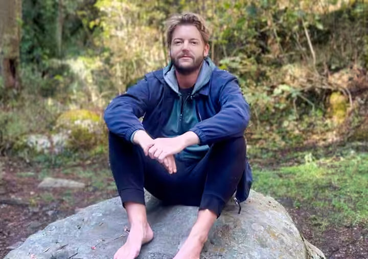A man sitting barefoot on a large rock in an outdoor forest setting, wearing a dark blue zip-up hoodie over a green shirt and dark pants.