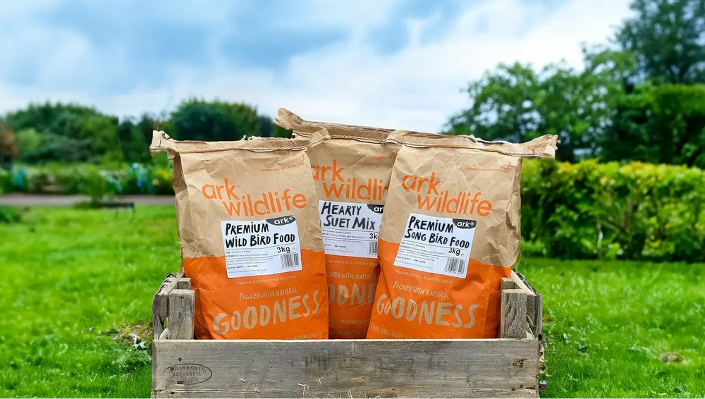 Three bags of Ark Wildlife Bird Food in a wooden crate on a green lawn with a garden background.